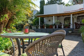 Eine Terrasse im Freien mit Korbstühlen und einem Tisch, umgeben von üppigem Grün und einem weißes Gebäude mit einem Ziegeldach.