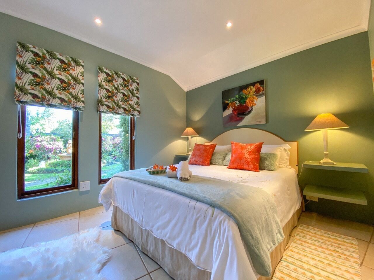 A bedroom with a large bed, two bedside lamps, a nightstand, and a view of a garden through the windows.