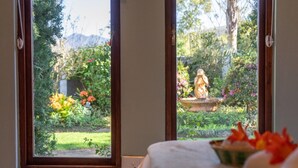 Une chambre avec vue sur un jardin, visible par une fenêtre, une statue sur un piédestal et un panier de fleurs sur le lit.