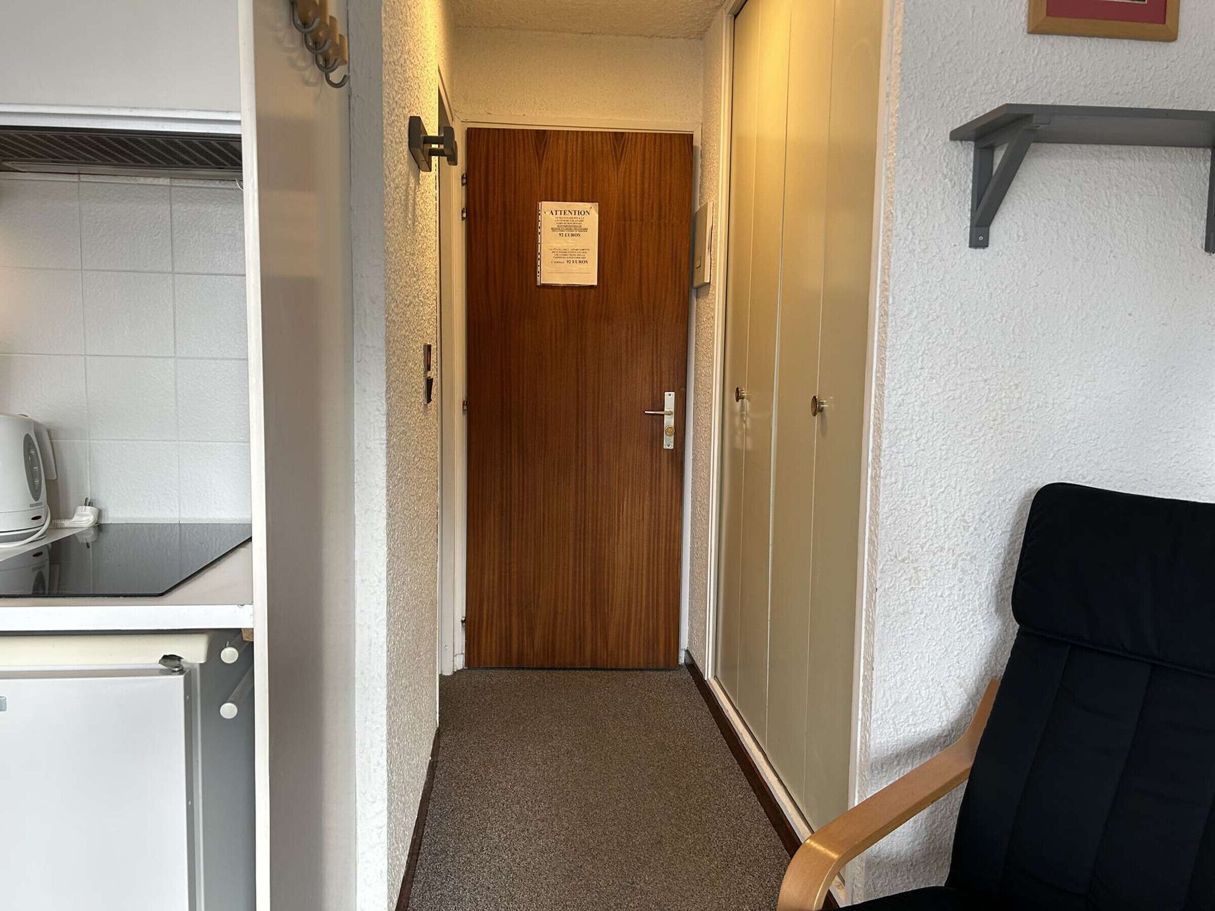 A narrow hallway with a wooden door at the end, a small kitchenette on the left, and a black chair on the right.