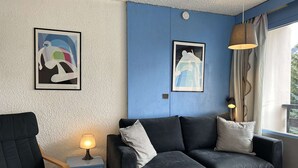 A room with a blue accent wall, a black sofa with cushions, a wooden chair, a small table with a lamp, and two framed abstract artworks.