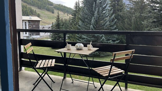 A balcony with a table and chairs, overlooking a mountainous landscape with trees and buildings.