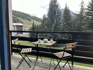 A balcony with a table and chairs, overlooking a mountainous landscape with trees and buildings. - Mountain Studio for 2 with West Balcony and Wi-Fi - Comfort in Montgenèvre (Montgenèvre)