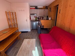 Un espacio habitable compacto con kitchenette, un banco de madera, un sofá rojo y un cesto de basura.