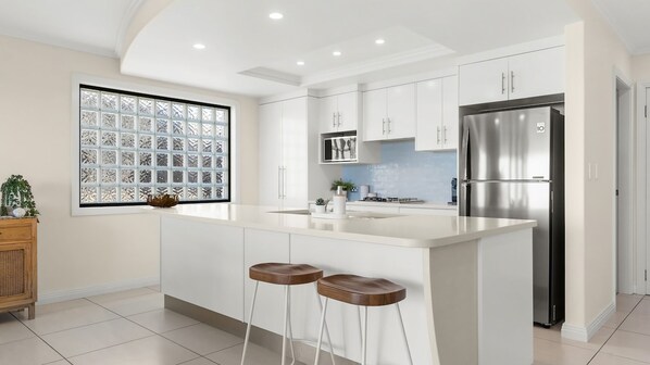 A modern kitchen with white cabinetry, a stainless steel refrigerator, and a large window with a geometric pattern. - Shawhaven - Shaws Bay Holiday Home (East Ballina)