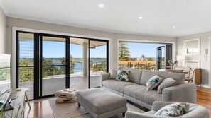 A spacious living room with a large grey sofa, a matching ottoman, and a view of a lake through large sliding glass doors. - Shawhaven - Shaws Bay Holiday Home (East Ballina)