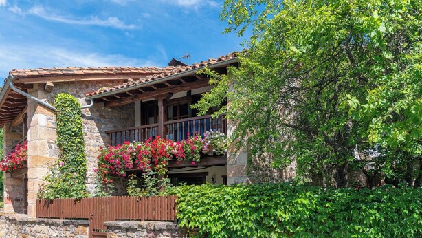 Un edificio de piedra con balcón de madera, rodeado de abundante vegetación y plantas en flor.