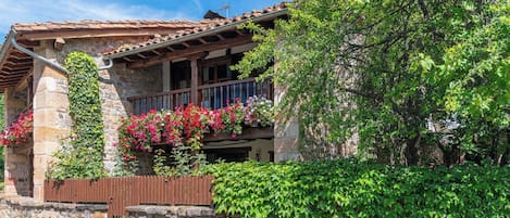 Un bâtiment en pierre doté d’un balcon en bois, entouré d’une végétation luxuriante et de plantes en fleurs.