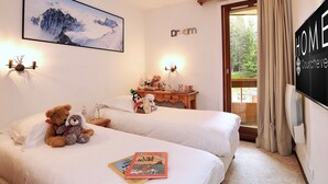 Quarto de hotel com duas camas, uma pintura de montanha, uma cômoda de madeira e uma TV exibindo a palavra 'HOME'.