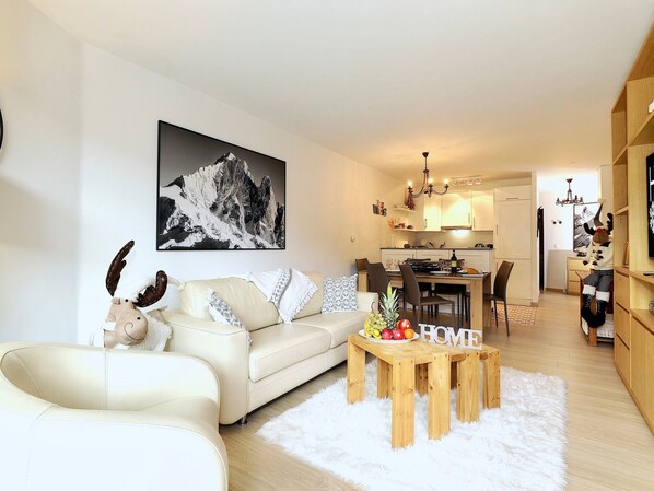 A modern living room with a sofa, a dining table with fruit, and a kitchen in the background.