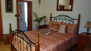 Un dormitorio con una cama, un cabecero de madera, una mesita de noche con una lámpara, un espejo y una puerta que da a otra habitación.