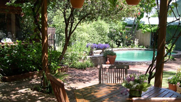 Banco y mesa de madera bajo una pérgola, rodeados de abundante vegetación y una piscina.