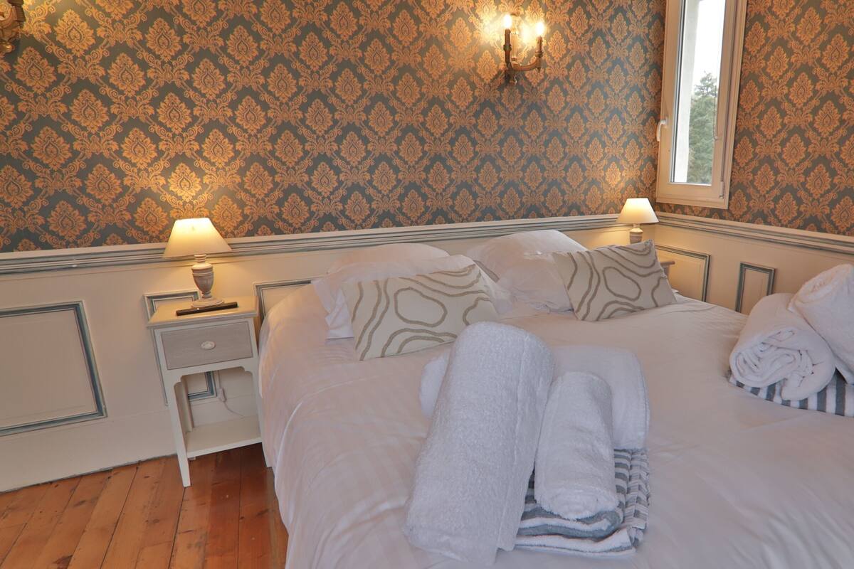 A neatly made bed with white linens and towels, two bedside lamps, a nightstand, and a window with a view.