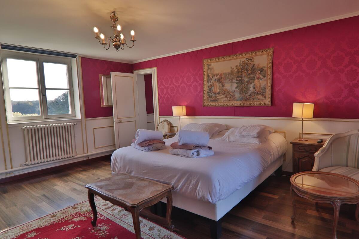 A bedroom with a bed, a chair, a table, a painting, and a window.