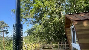 Una terraza de madera con sauna, una construcción de madera con techo inclinado y una estructura metálica con parte superior circular.