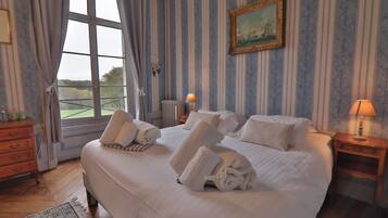 A neatly made bed with towels, a wooden dresser, a nightstand with a lamp, a painting on the wall, and a window with curtains.