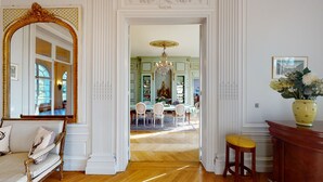 Una habitación con un espejo grande y decorativo, un comedor con una araña y un jarrón con flores sobre una mesita auxiliar.