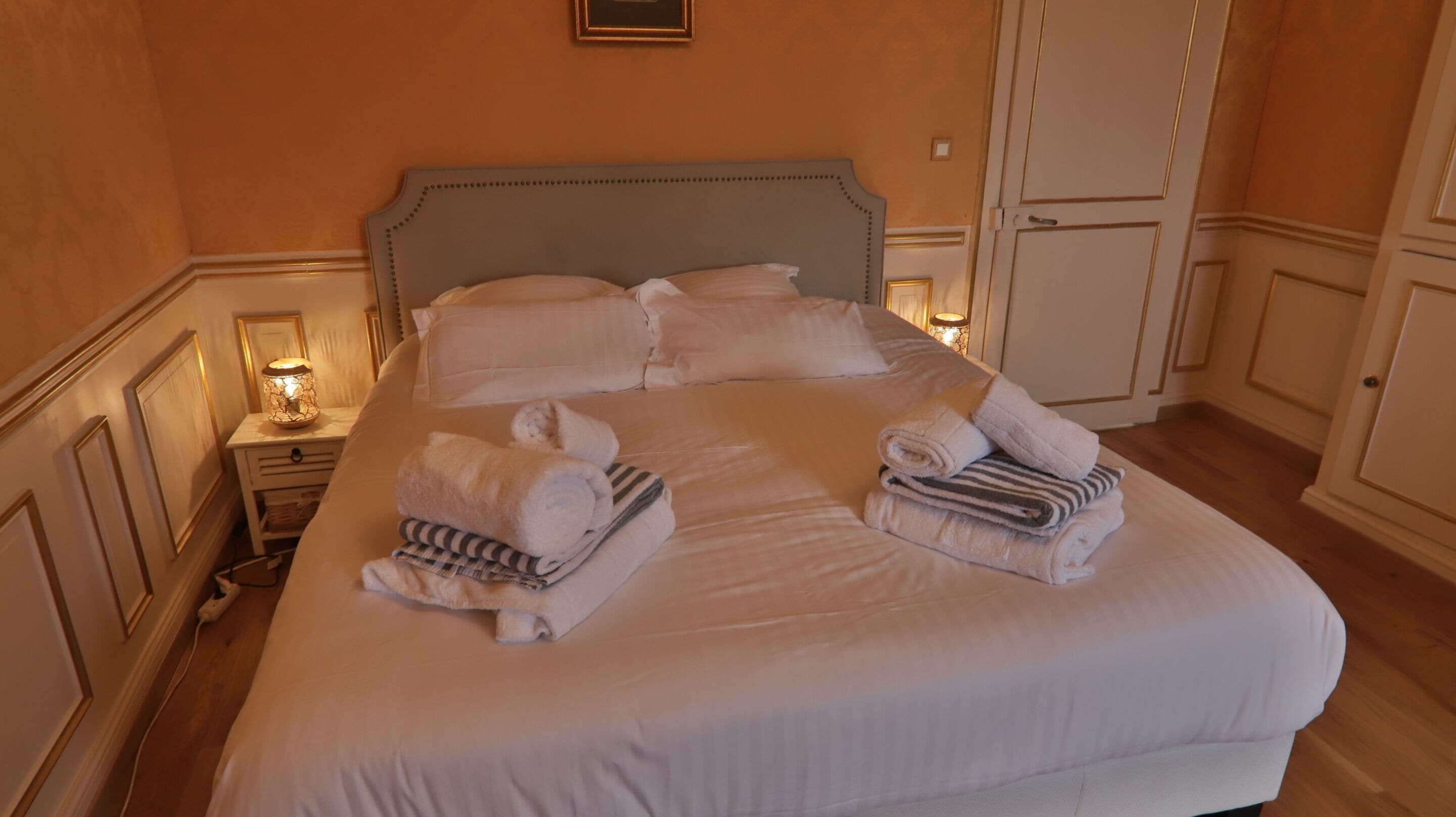 A neatly made bed with pillows and folded towels, a bedside lamp, and a wardrobe in a hotel room.