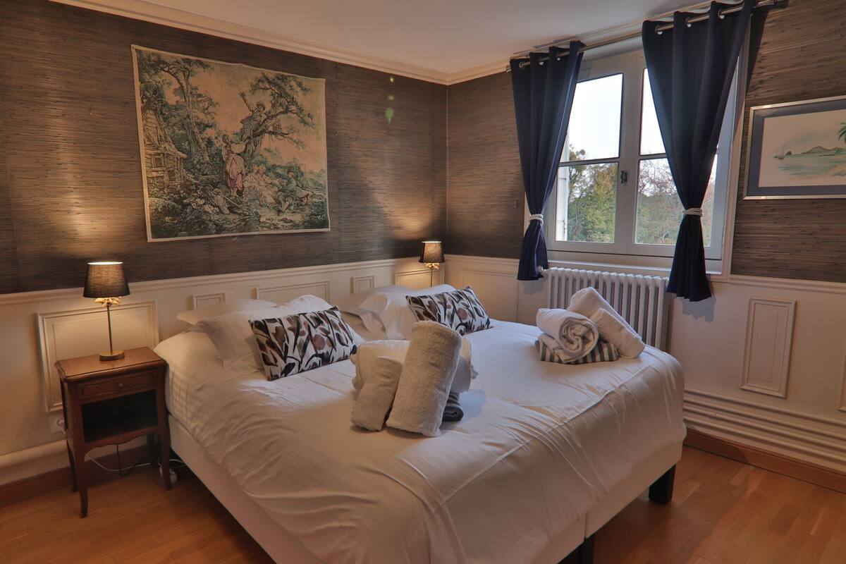 A hotel room with a large bed, two bedside tables, a window with curtains, and a painting on the wall.
