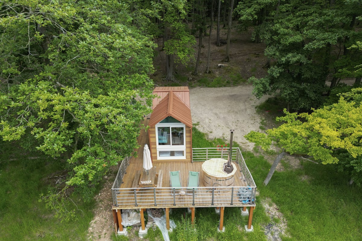 A wooden cabin with a deck, a hot tub, and a view of the surrounding forest.