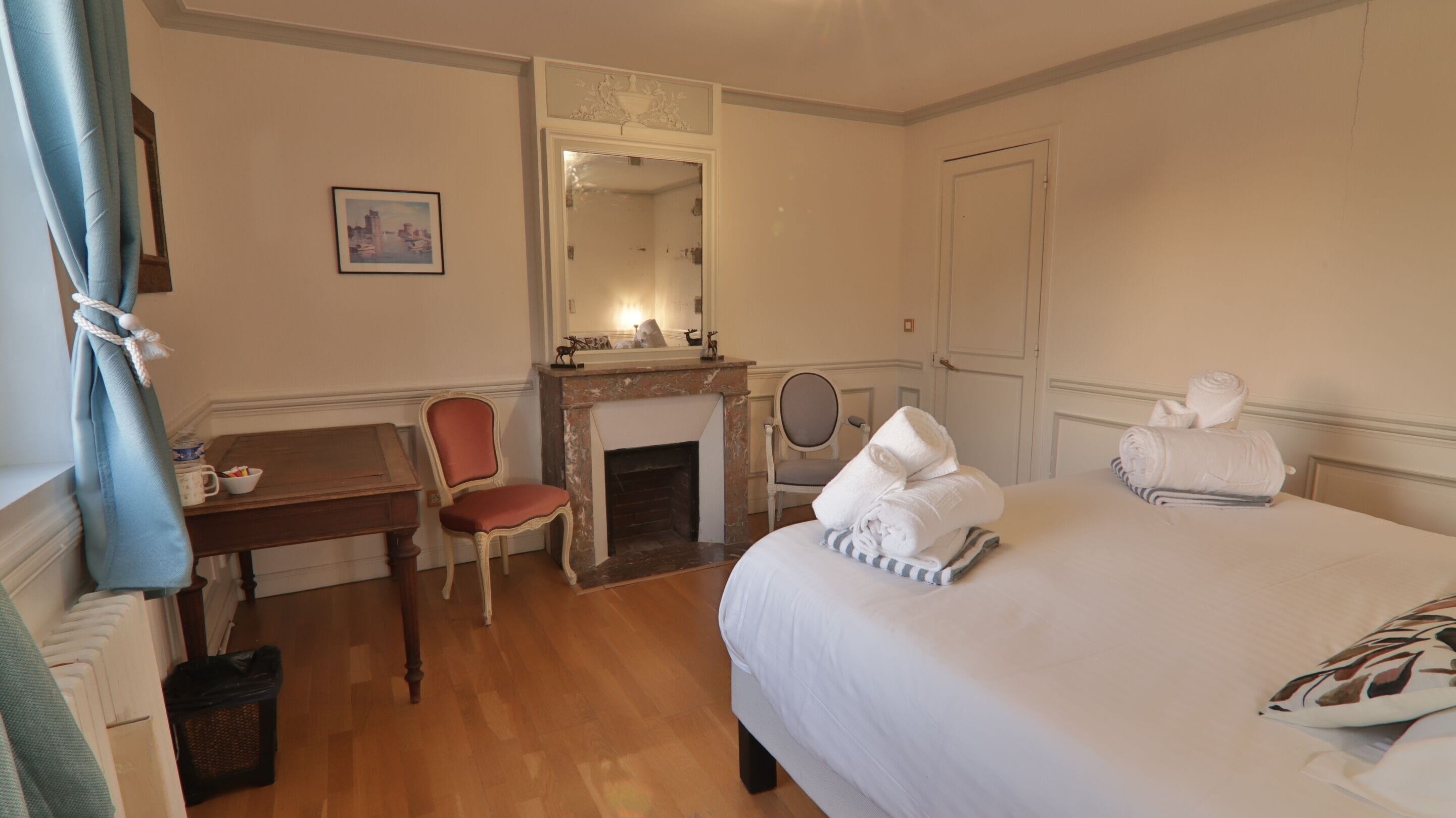 A hotel room with a bed, a desk, a chair, a fireplace, a mirror, and a door.