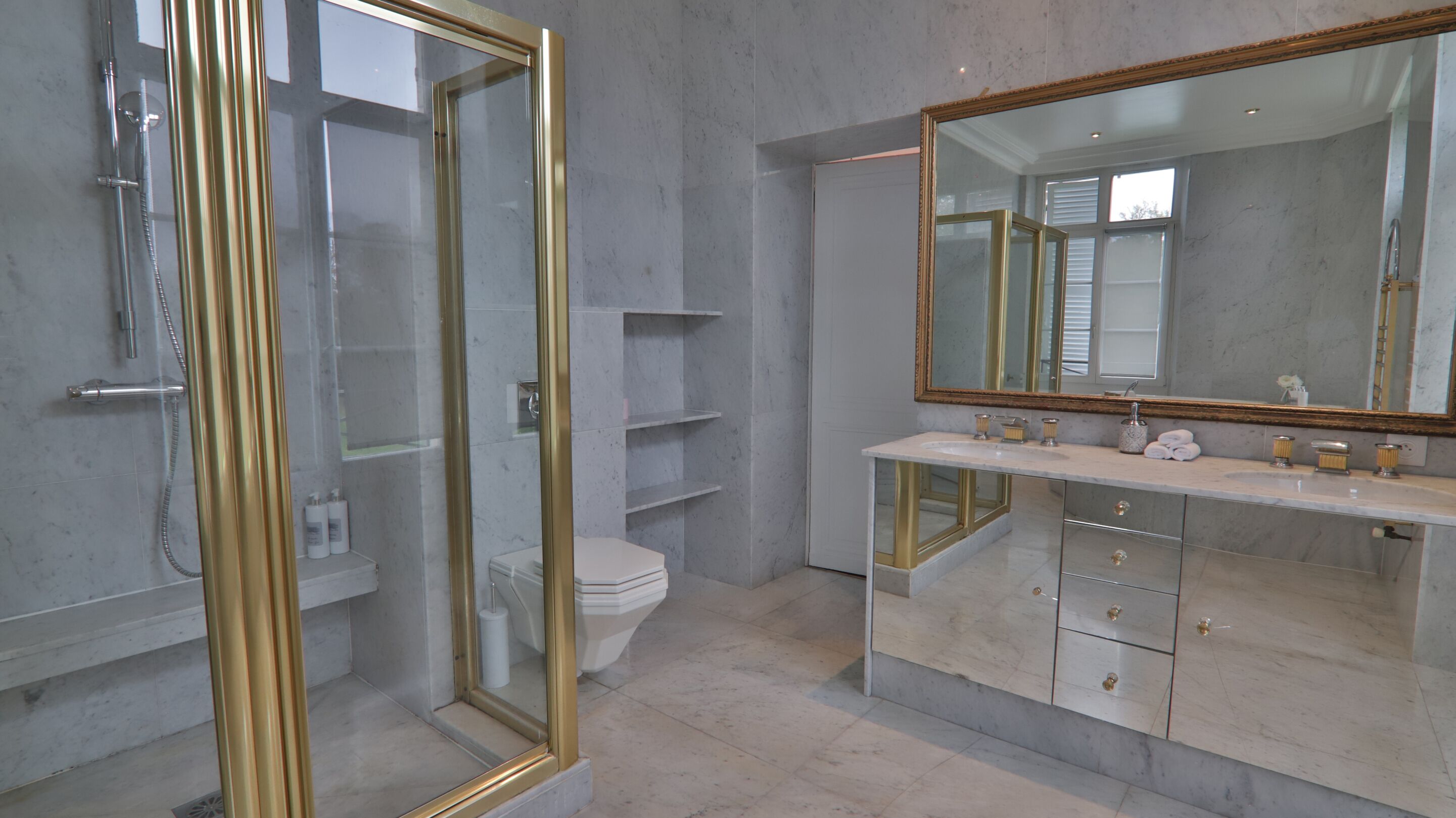 Un baño con cabina de ducha de vidrio, inodoro, tocador con espejo grande y suelo de mármol.