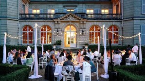 Un área de comedor al aire libre con mesas y sillas, rodeada por un edificio con ventanas arqueadas y un balcón.