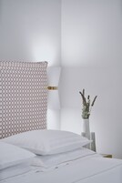 Una cama bien hecha con sábanas blancas, una almohada estampada y un florero con flores secas sobre una mesita de noche.
