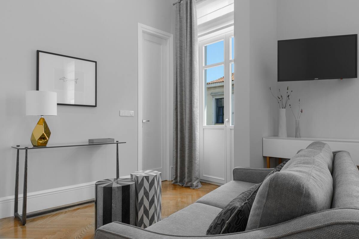 Ein modernes Wohnzimmer mit grauem Sofa, einem Beistelltisch mit Lampe, einem gerahmten Bild an der Wand und einem Flachbildfernseher, der über einem weißen Konsolentisch angebracht ist.