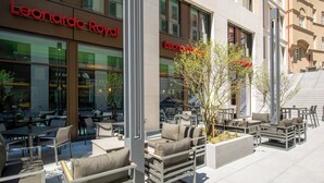An outdoor seating area with tables and chairs, a small tree, and a building with a sign that reads "Leonardo Royal". - Leonardo Royal Hotel Nürnberg (Nuremberg)