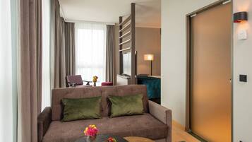 Una habitación de hotel moderna con un sofá marrón, cojines verdes, una pequeña mesa redonda con frutero y flores, un escritorio con una silla y una puerta corrediza.