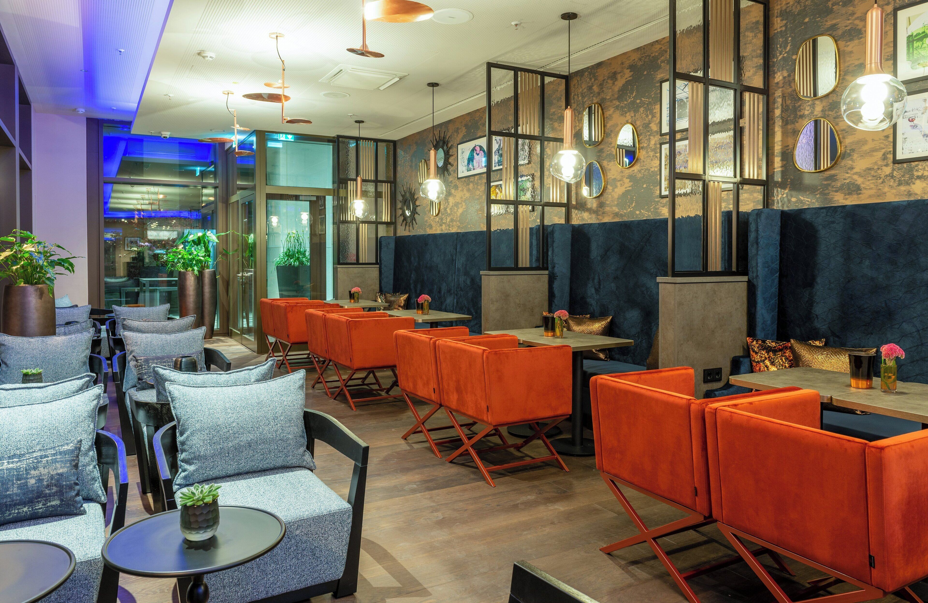 Interior de un restaurante moderno con mesas altas y asientos acolchados, espejos decorativos y plantas en macetas.