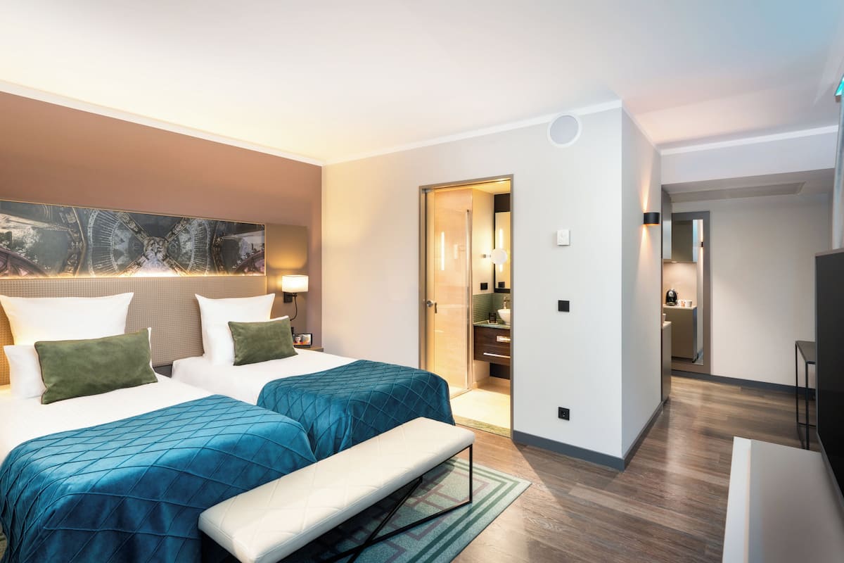 Quarto de hotel com duas camas, um banco, uma grande obra de arte na parede e banheiro visível através de uma porta aberta.