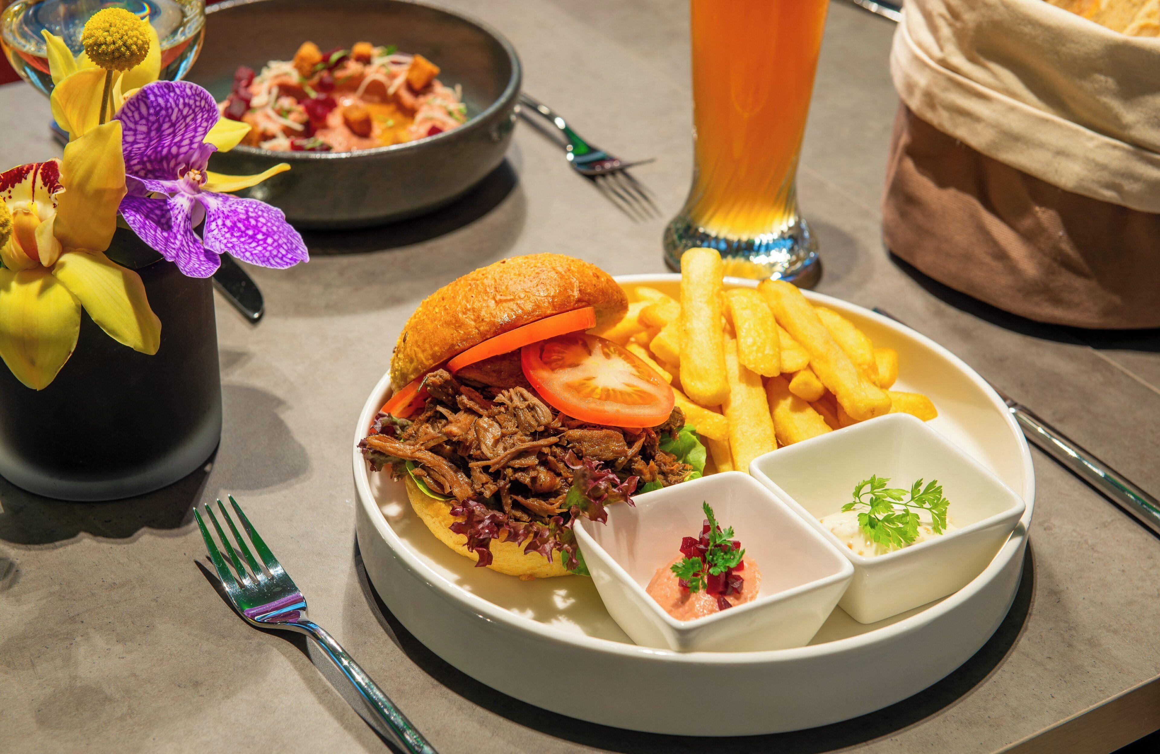 A plate with a sandwich, fries, and two dipping sauces, accompanied by a glass of beer and a bowl of salad.