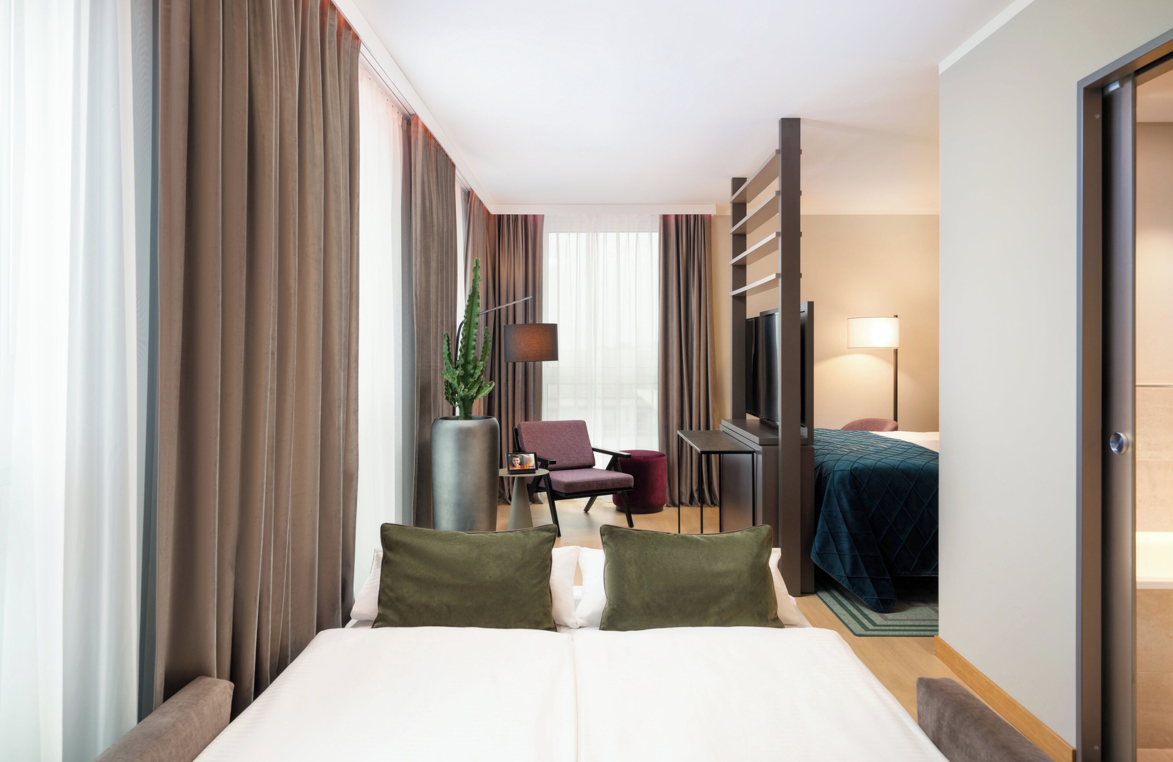 A hotel room with a large bed, a desk, a chair, a TV, and a plant.