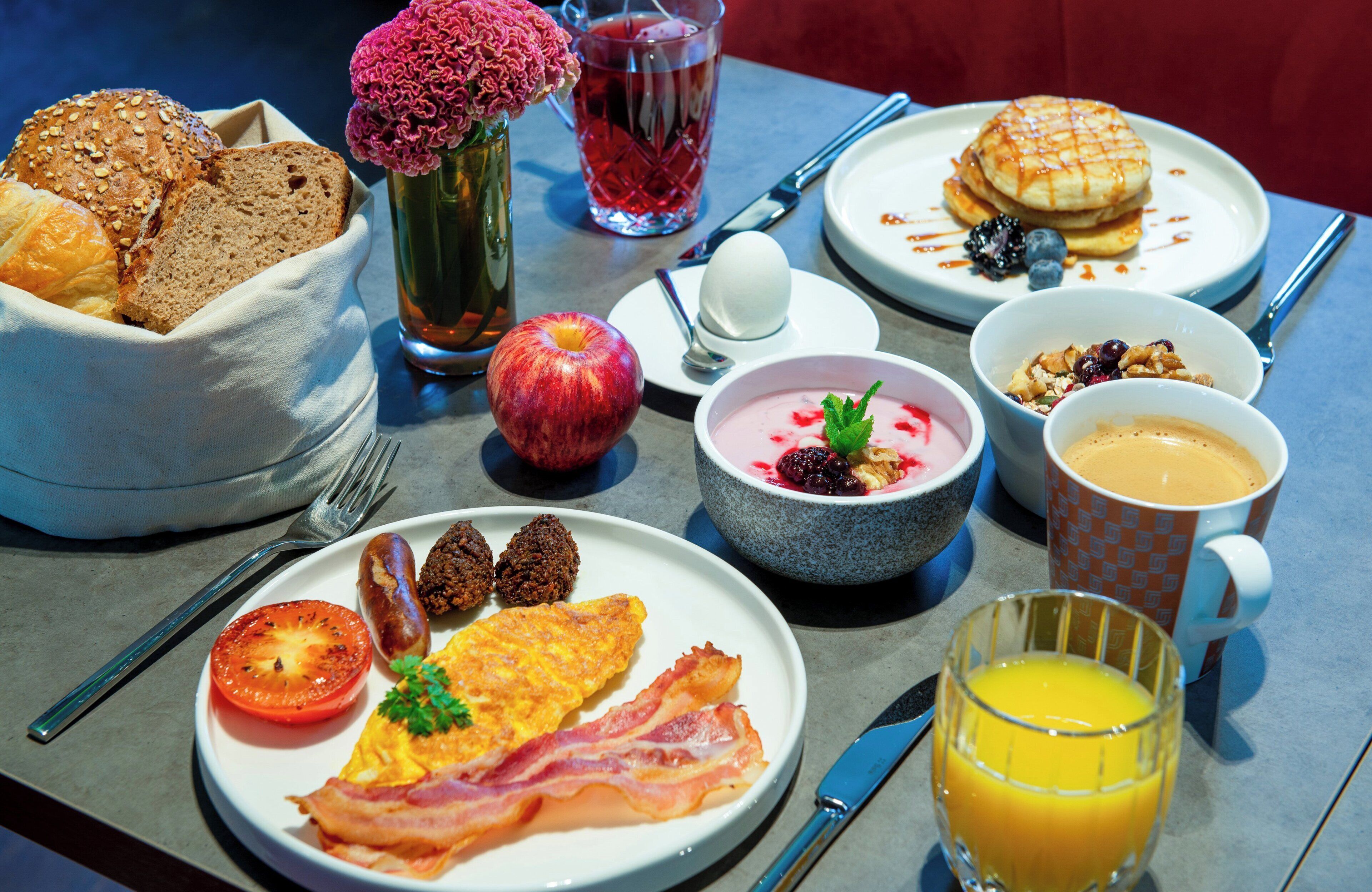 Una mesa de desayuno con diversas opciones de comida, entre ellas pan, huevos, tocino y un vaso de zumo de naranja.
