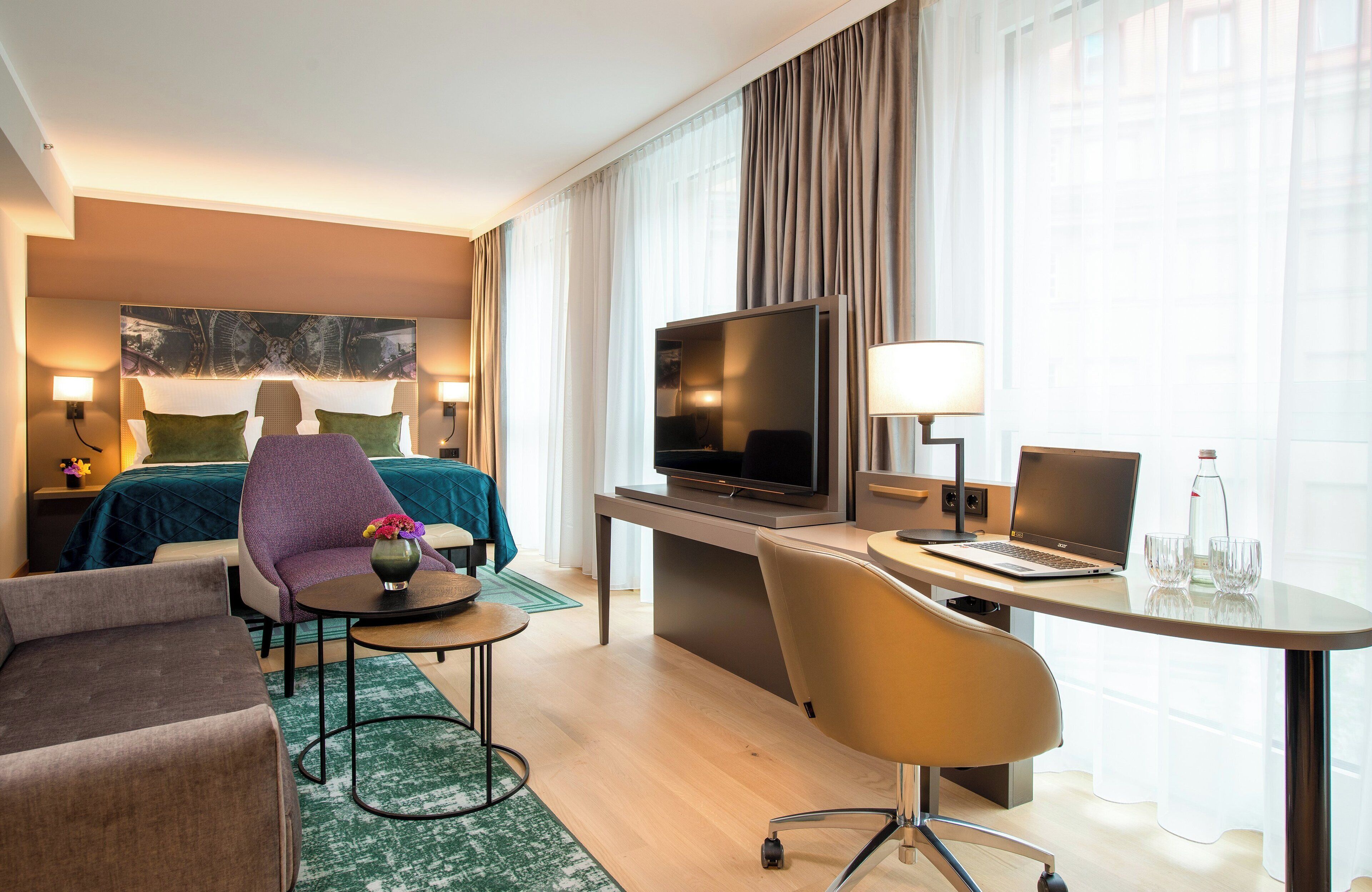Ein modernes Hotelzimmer mit einem Bett, einem Schreibtisch mit Laptop, einem Fernseher und einer Sitzecke mit Sessel und kleinem Tisch.