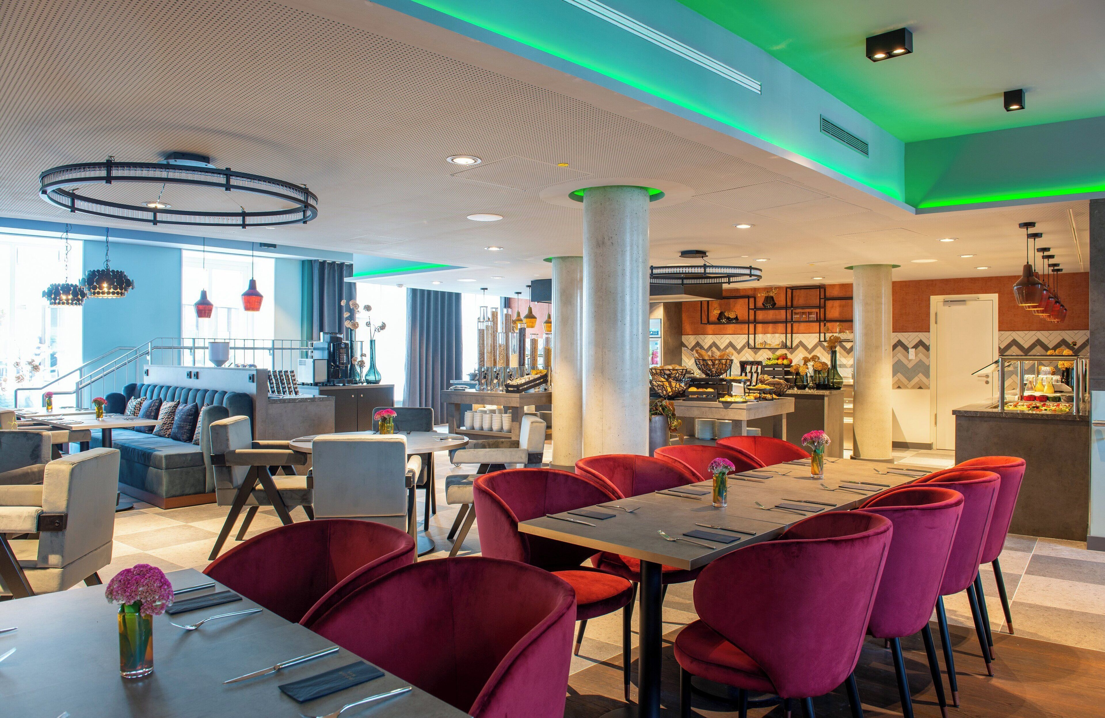 A modern dining area with red chairs, a long table, and a bar area in the background.