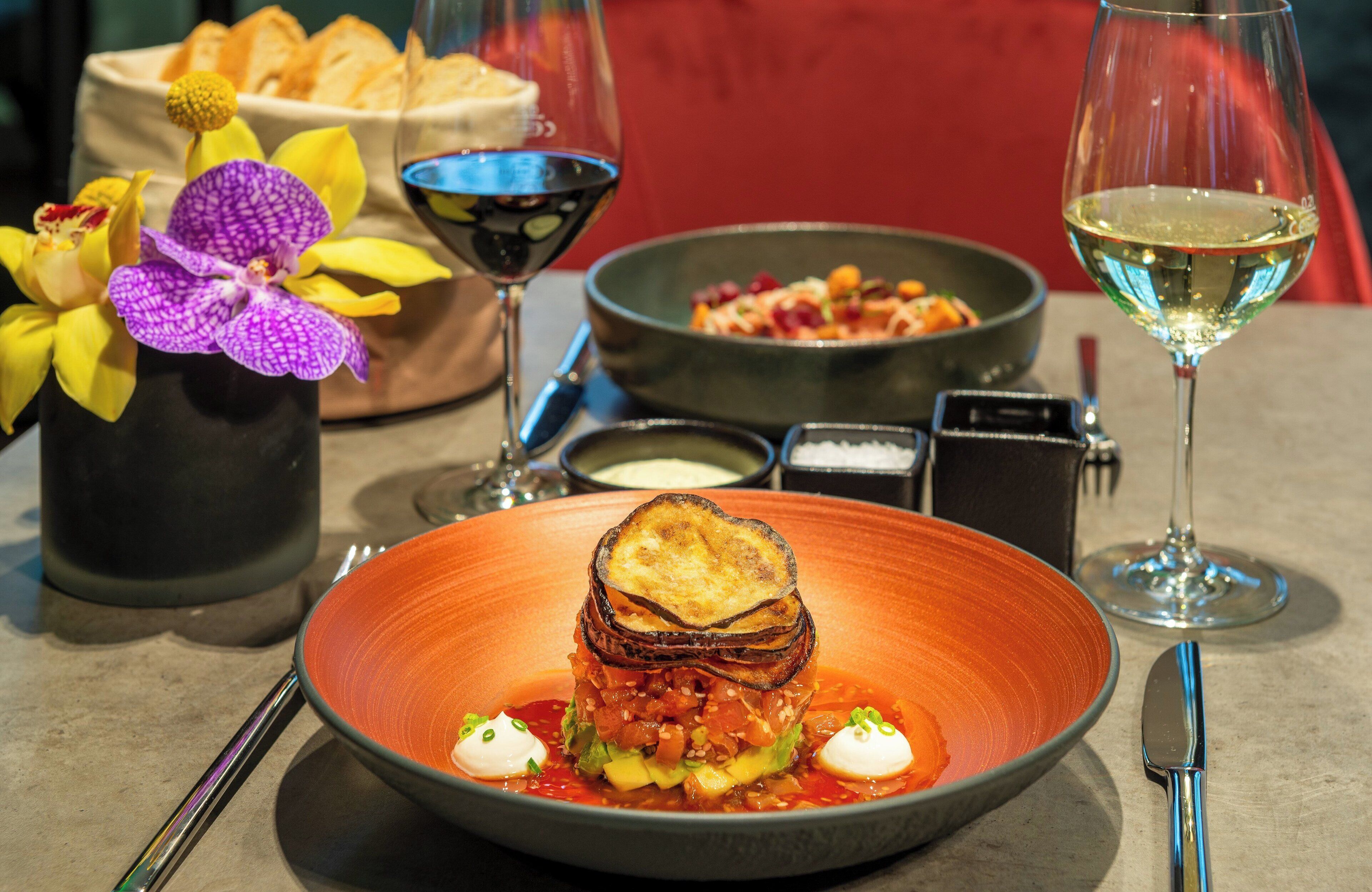 A dining table with a plate of food, two wine glasses, a vase with flowers, and a bowl of soup.