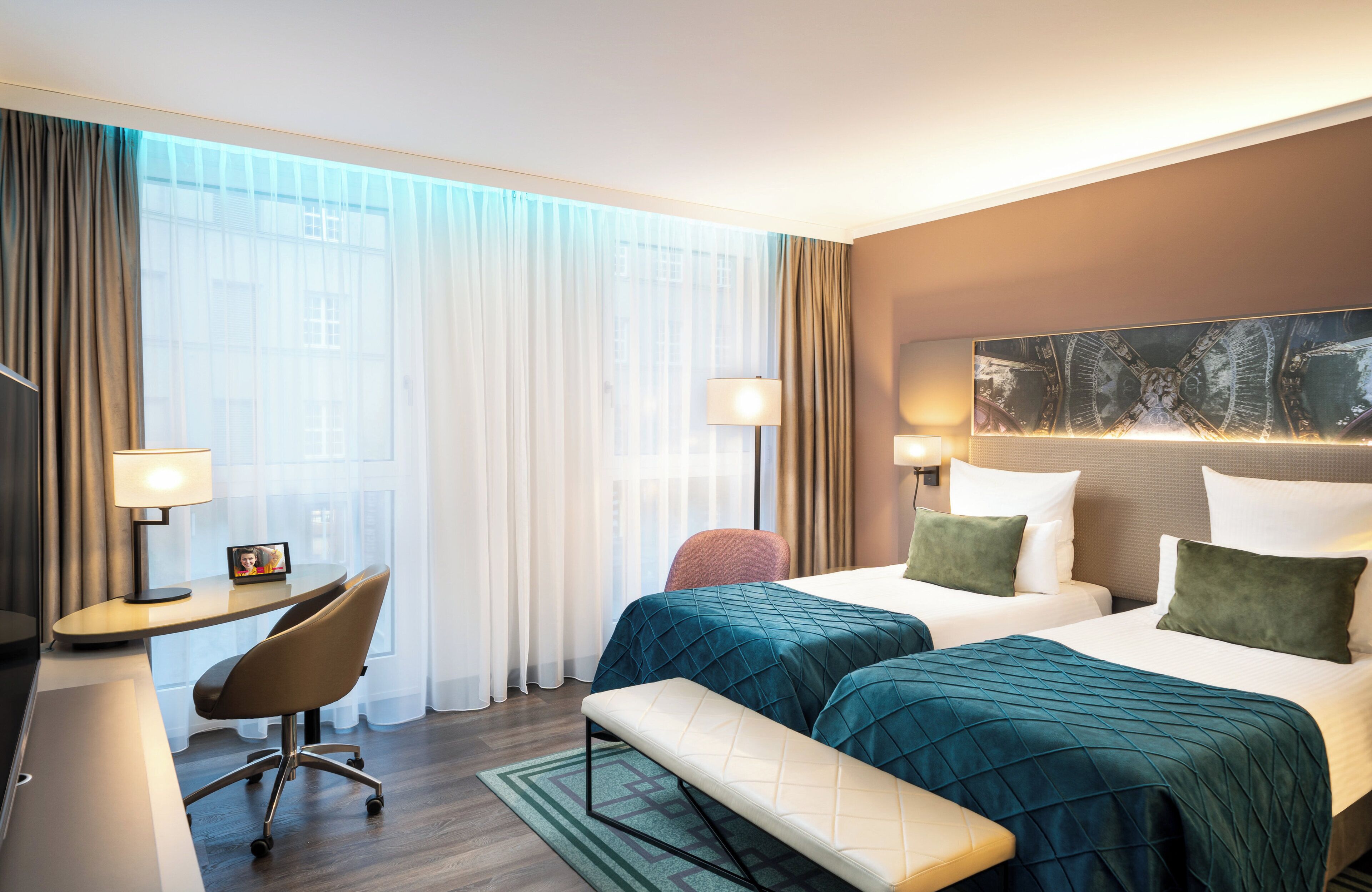 Una habitación de hotel con dos camas, un escritorio, una silla, un banco y un ventanal con cortinas.