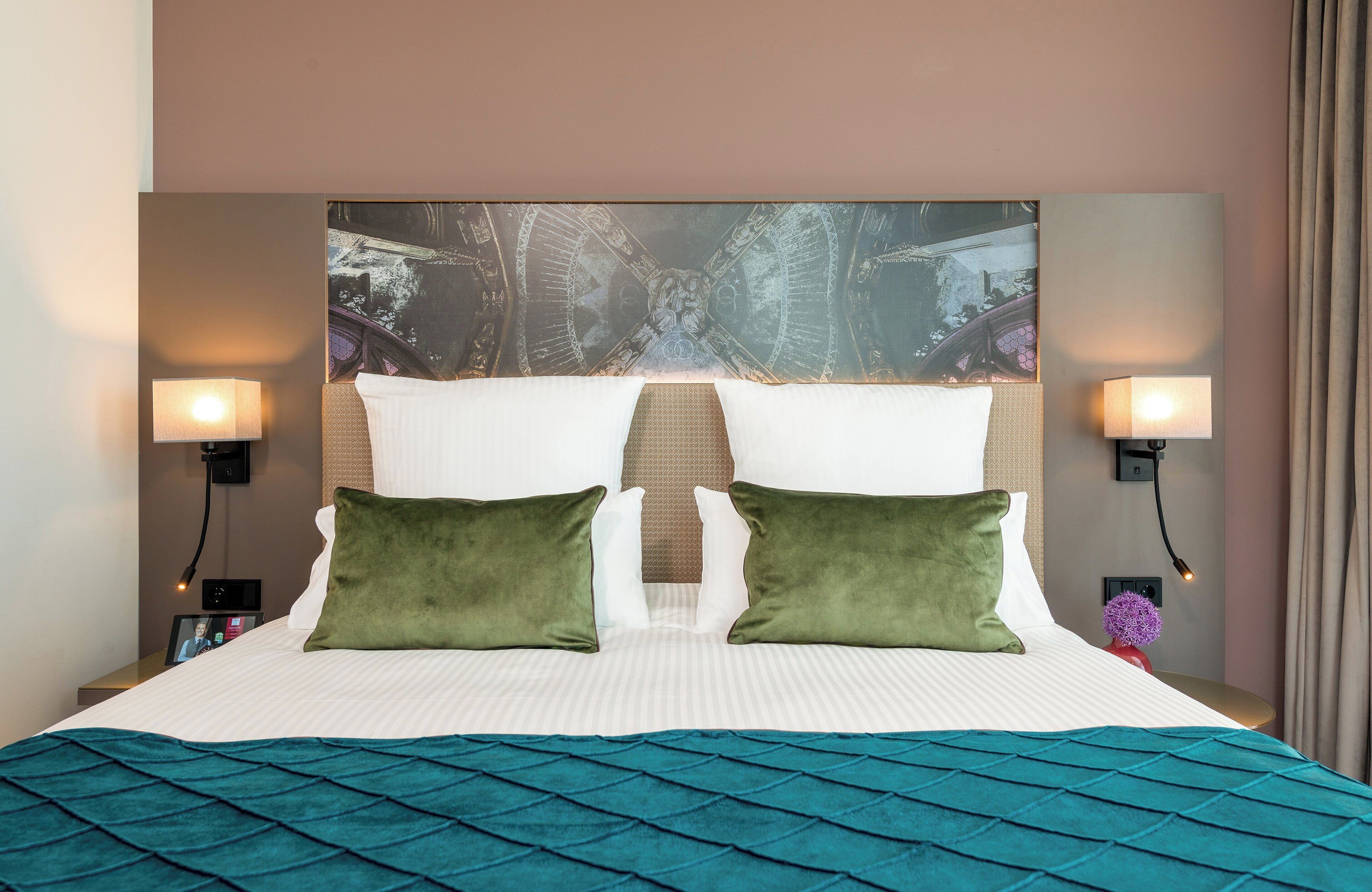 A hotel room with a large bed, two green pillows, a headboard with a decorative panel, two wall-mounted lamps, and a nightstand with a photo frame.