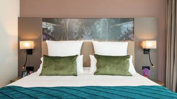 A hotel room with a large bed, two green pillows, a headboard with a decorative panel, two wall-mounted lamps, and a nightstand with a photo frame.