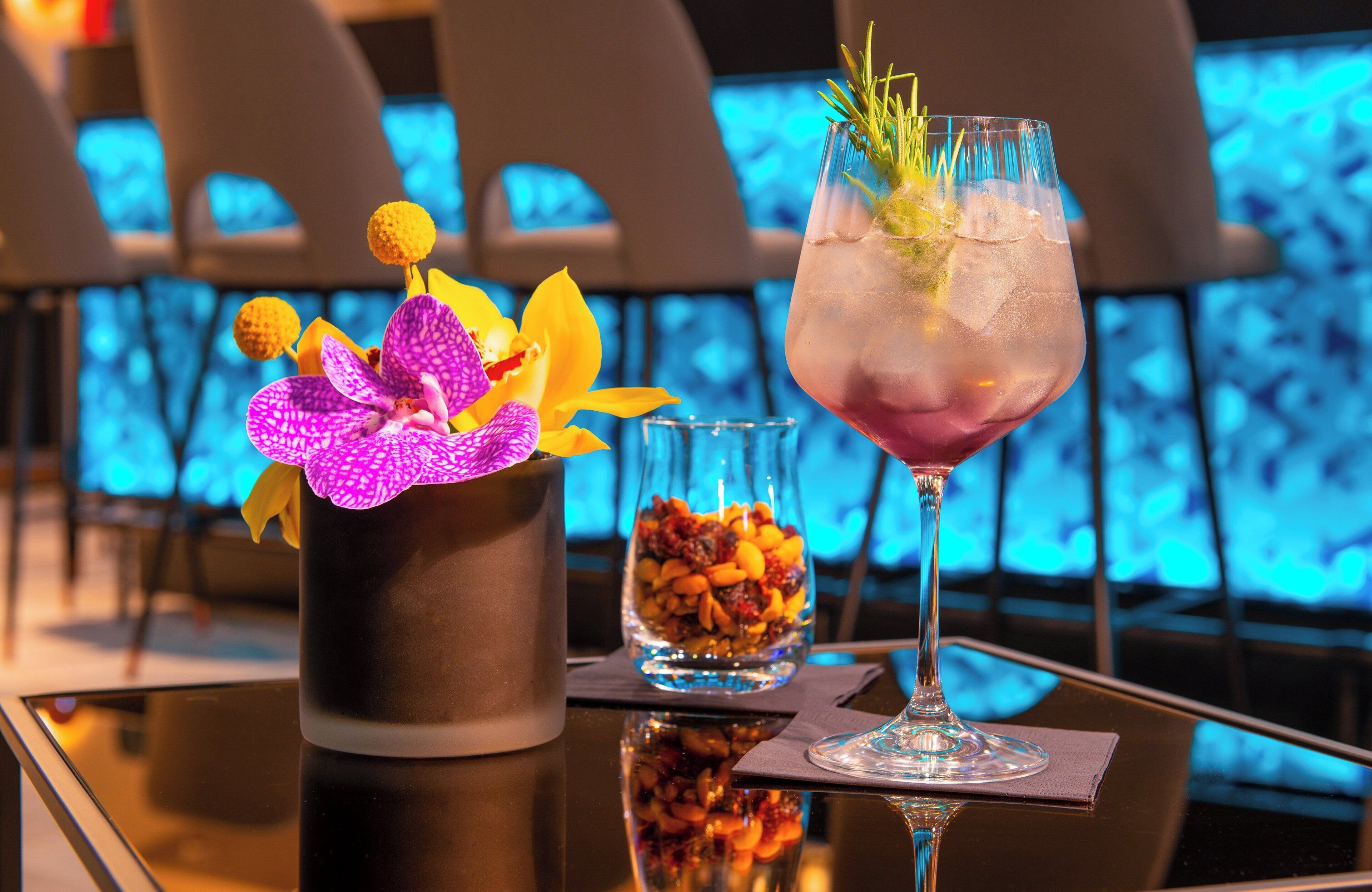 A table with a glass of iced cocktail garnished with a sprig of rosemary, a small glass of nuts, and a purple orchid in a black pot.