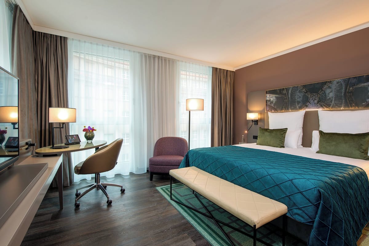 Quarto de hotel com uma cama grande, uma escrivaninha com cadeira, um laptop, um vaso com flores e uma janela ampla com cortinas.