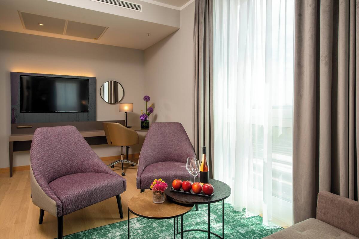 Quarto de hotel moderno com televisão de tela plana, duas poltronas roxas, uma pequena mesa redonda com frutas e copos, e uma grande janela com cortinas.