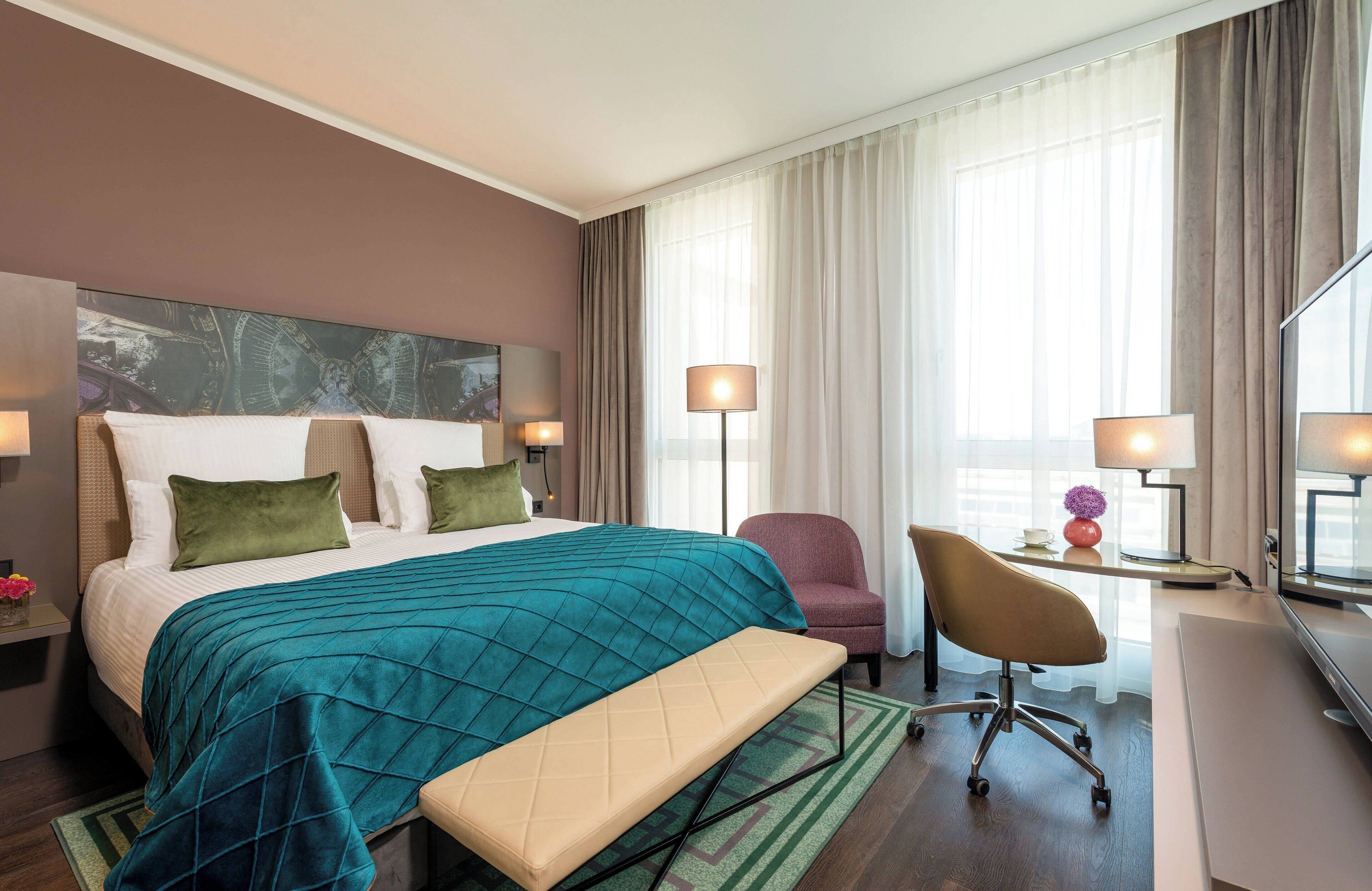 Quarto de hotel com uma cama grande, uma escrivaninha com cadeira, uma luminária e vista para a cidade pela janela.