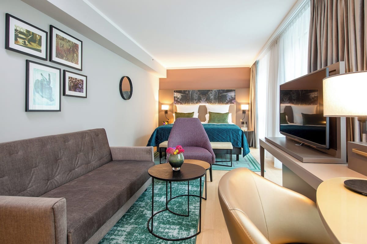 Quarto de hotel com sofá, mesa redonda pequena com um vaso, cama com cabeceira azul, televisão e quadros nas paredes.