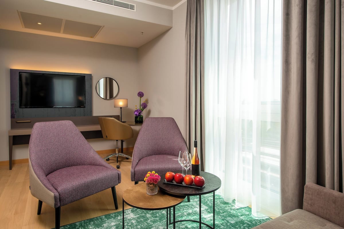 Une chambre d’hôtel moderne équipée d’un téléviseur à écran plat, de deux fauteuils violets, d’une petite table ronde avec des fruits et des verres, et d’une grande fenêtre avec des rideaux.
