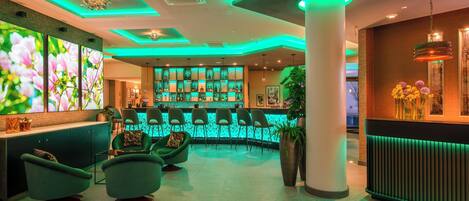 A modern bar area with green ambient lighting, a large screen displaying floral images, and a seating area with green chairs.