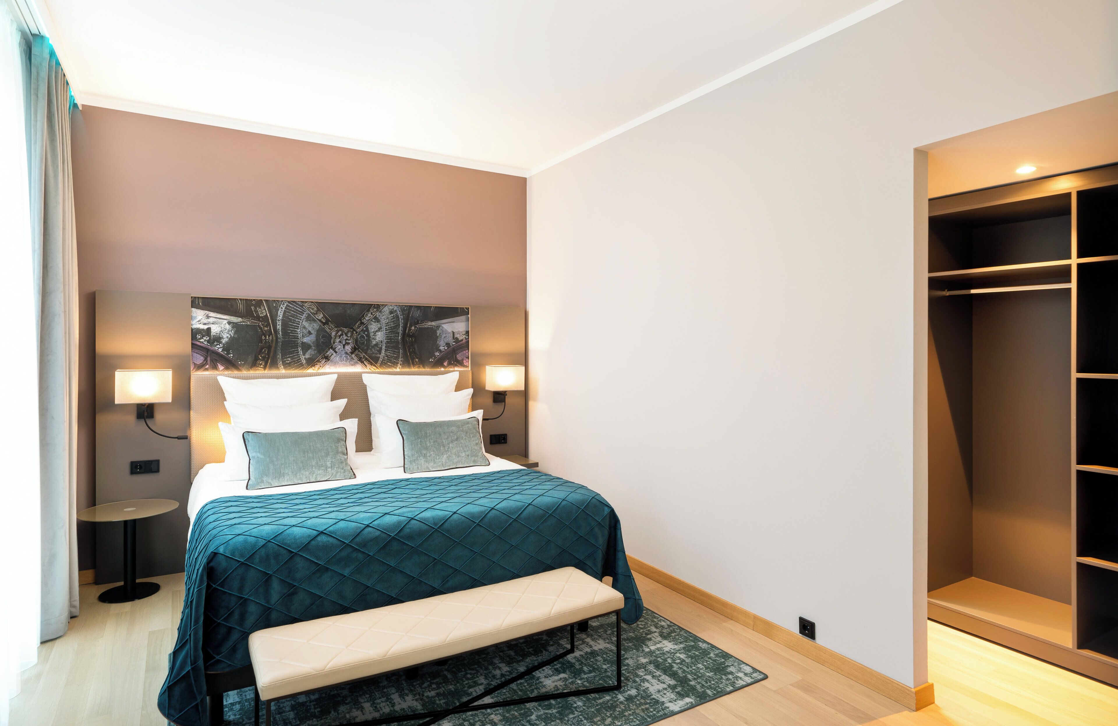 A modern hotel room with a large bed, bedside lamps, a bench, and a walk-in closet.